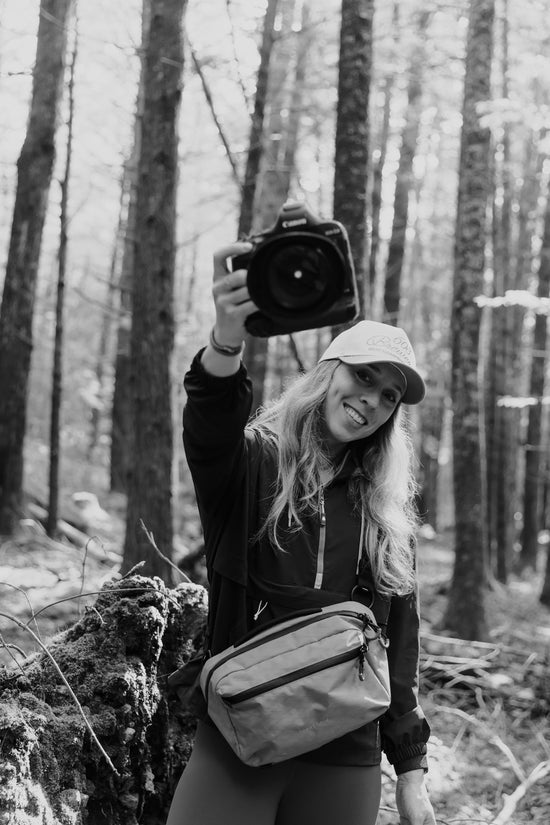 A photographer in a forest wearing a Kiboko City Sling 6L at her waist and holding up a Canon camera, black and white
