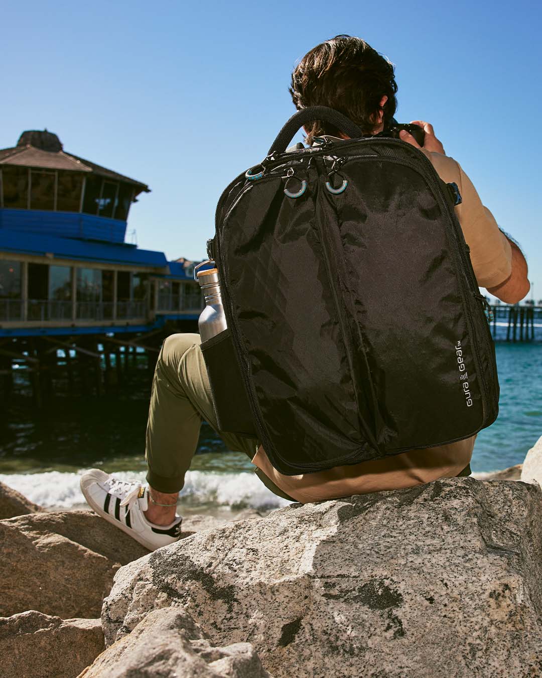 A man sitting on rocks by the sea wearing a black Kiboko 2.0 22L+