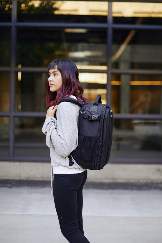 A woman wearing a black Kiboko 2.0 22L+ on her back, side view in front of a glass building