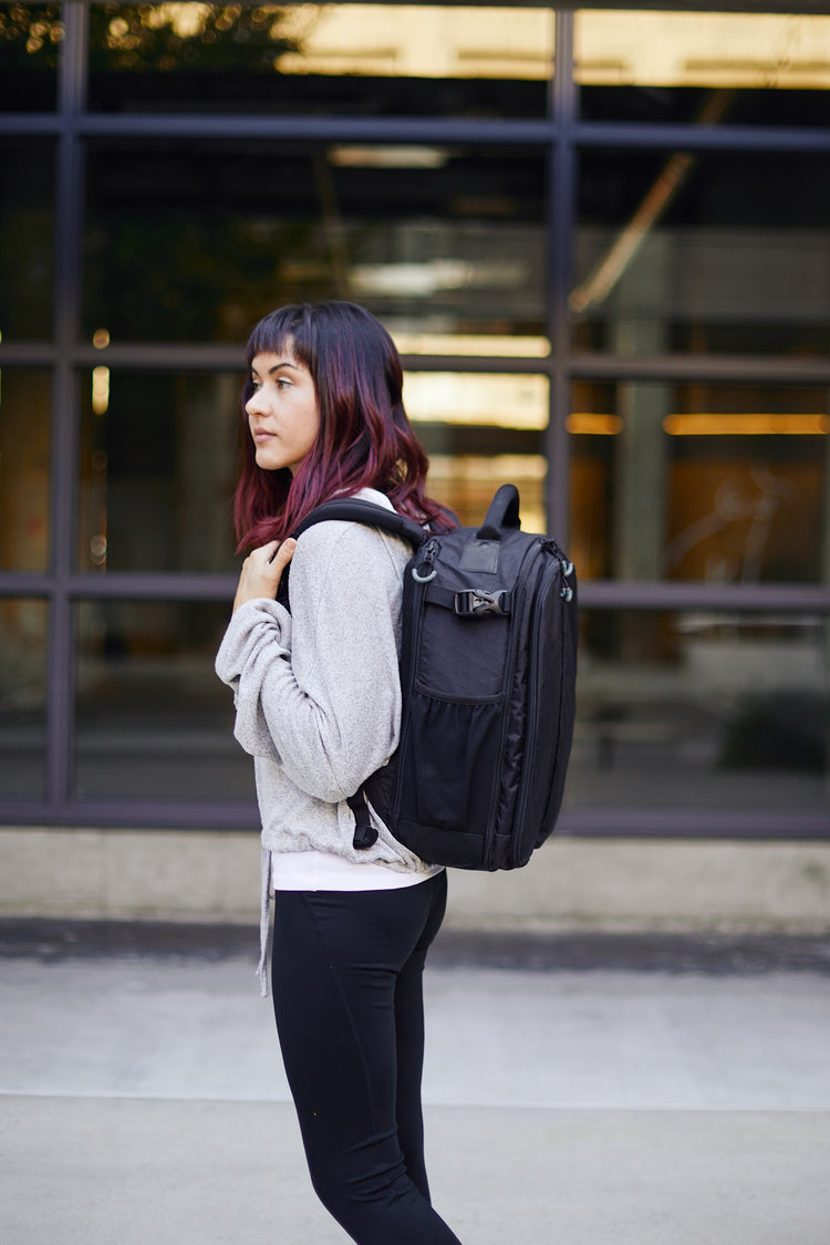 A woman wearing a black Kiboko 2.0 22L+ on her back, side view in front of a glass building