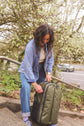 A woman unzipping a Forest Green Kiboko 2.0 30L+ on outdoor stairs